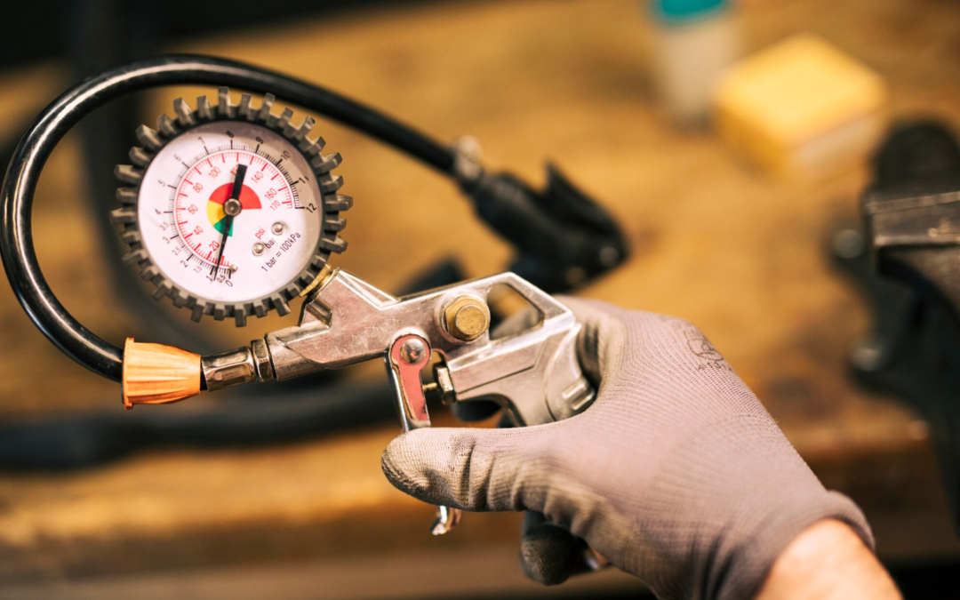 Uma imagem colorida, em close-up, mostra uma pessoa usando uma luva de trabalho texturizada cinza, segurando uma pistola de inflador pneumático manual e um medidor de pressão. A pessoa segura a pistola de inflador de metal escovado, com seu gatilho de operação, na mão direita. O medidor de pressão circular está montado em cima da pistola, conectado por uma mangueira curta de borracha preta. O medidor tem uma moldura de borracha texturizada preta e um mostrador branco com marcas vermelhas e pretas. O ponteiro vermelho do medidor indica uma leitura de cerca de 22 PSI (ou 1,5 bar), o que sugere um estado de baixa pressão, relevante para o tópico do texto sobre diagnóstico de falhas em compressores de ar. A parte inferior da pistola de inflador tem um pequeno acoplamento de metal. No fundo, que está desfocado (bokeh), há uma bancada de trabalho de madeira escura com ferramentas e equipamentos desfocados, incluindo uma morsa de bancada e um recipiente de óleo ou graxa, contextualizando a cena como um ambiente de oficina industrial ou de reparo. A iluminação é natural e difusa, focando na mão e no equipamento pneumático.